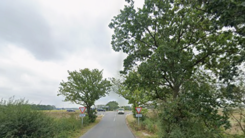 Part of a country road has trees and bushes on both sides. There are give way signs as a main road lies ahead. There is a van on the small road and two cars can be seen on the main road in the distance.