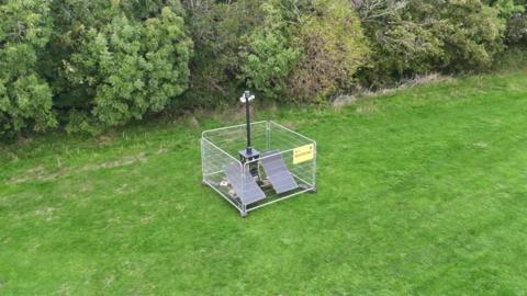A large pole with speakers and CCTV cameras at the top. At the foot of the pole are two solar panels, surrounded by fencing. It is placed on a green field with trees in the back.