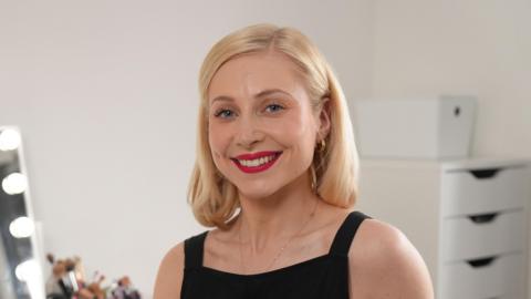 Amy, who has shoulder length blonde hair and wears a sleeveless black top, smiles at the camera. Behind her is a mirror and a number of makeup brushes