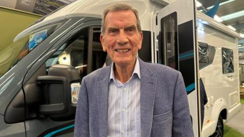 A man in a pale blue pinstripe shirt and blue suit jacket standing in front of a white and blue motorhome. He is smiling. 