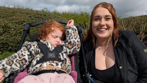 A little girl sitting in a pram. Her mother is smiling at the camera next to her. 