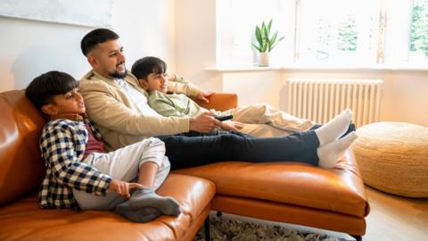 A father sits on a leather sofa with his two young sons either side. He is smiling and holding a remote control.