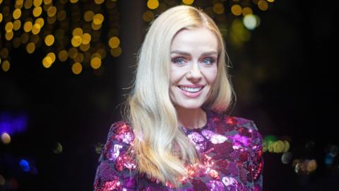 Katherine Jenkins wearing pink sequinned top and smiling at the camera. She has long blonde hair worn down and the backdrop to the photo is dark, with some twinkly lighting