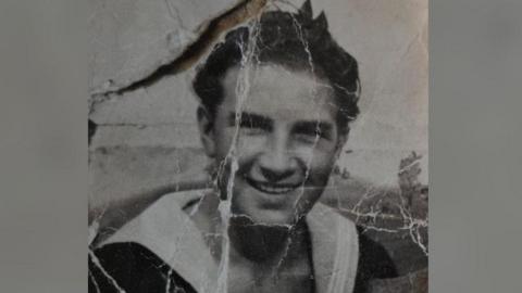 A black and white picture of a young man, He has short dark hair and is looking towards the camera, smiling. The photograph has white lines or creases on it.
