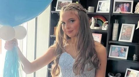 Maisie, pictured in a blue dress, holding balloons in front of a black display chest featuring family photos and ornaments