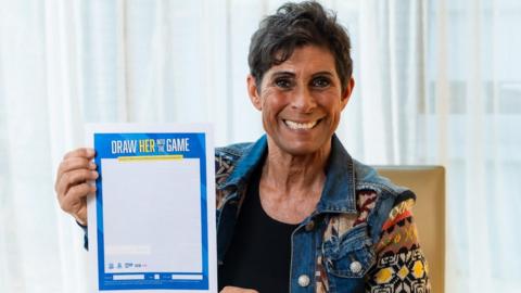 Fatima Whitbread with a grin on her face, holding up a sheet of paper that says Draw Her Into The Game