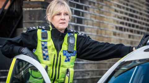 Sarah Lancashire as Sgt Catherine Cawood in Happy Valley, wearing a hi-vis police vest and leaning on a police car