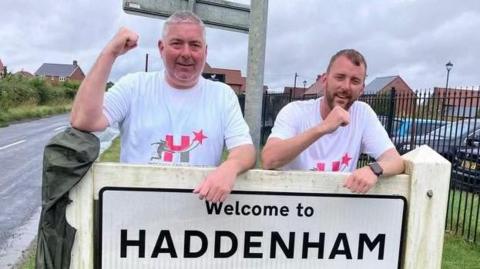 Two men leaning on Haddenham sign