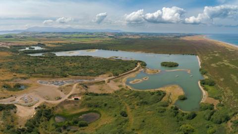 An ariel view of a nature reserve, there is a large pond in the middle, you can see the coastline on the other side of the reserve, on a clear blue day.