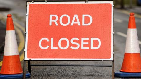 Close up of red road closed sign. Traffic cones are on either side of the sign.