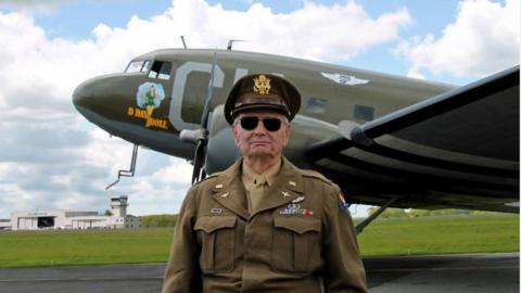 Lieutenant Colonel David Hamilton in military uniform standing in front of a military plane on a runway. He is wearing dark sunglasses