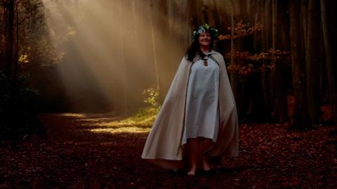 Deborah Rose Hālani stands barefoot in a wooded area wearing white robes. Rays of sunlight shine through the cracks in the trees illuminating the leafy ground underfoot.