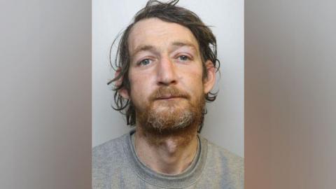 A police image of Ian McNally who has a rough beard and is wearing a grey t-shirt