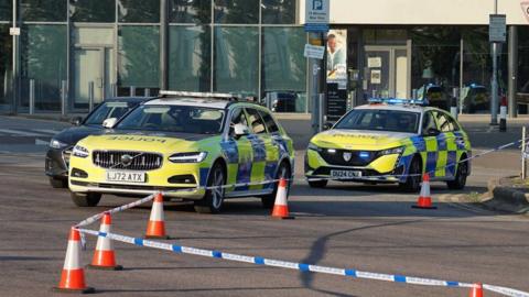 Police vehicles block off a section of road outside a modern glass-fronted building. Marked police cars are parked behind blue-and-white police tape and a line of orange traffic cones. Road signs, bollards and parked vehicles are visible around the cordon.