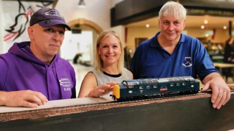 Karl Stevens, director KMS Railtech, who will install the railway, with Depute Provost Jackie Hendry and Graeme Swanson, chairman of Inverness & District Model Railway Club. They are standing inside the food hall and have a scale-model diesel locomotive in front of them.