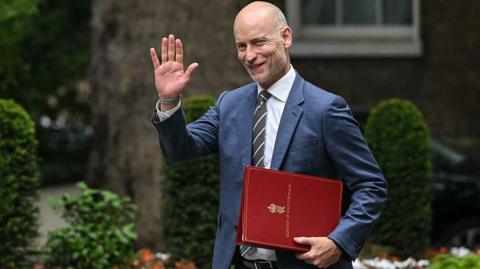Stephen Kinnock arrives at Downing Street carrying a red Welsh secretary file and raising a hand in greeting. Trees, trimmed shrubs and flowerbeds can be seen in the background.