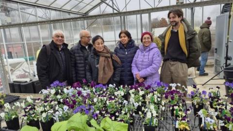 Six people standing in a glasshouse, with an shelf of potted flowers in front of them