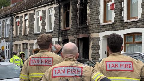 A stone terraced house with fire damage around the windows and doors. Three fire officers look at the house with their backs to the camera. 