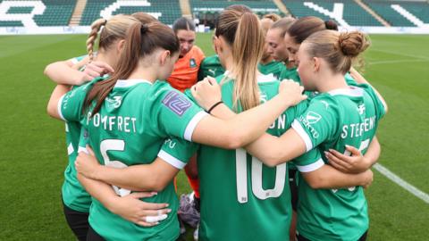 Nine women wearing Plymouth Argle's green football kits are standing in a circle on a football pitch facing each other with their arms wrapped round each other.