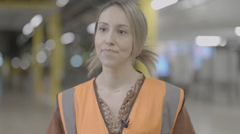 Adriana Genes has fair hair, parted in the middle and tied back. She is wearing a terracotta-coloured top and an orange hi-vis vest.