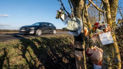 Flowers and cards at the site of the crash. A car drives past on the road above. It is a rural road and the sky is blue.