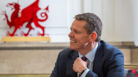 Rhun a Iorwerth sitting down, wearing a dark suit sits at a table, with a large red Welsh dragon emblem visible in the background of a light-coloured room.