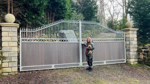 Tow gates, each 10ft wide, stand between two stone pillars. A man, half the height of the gates, stands beside them holding a small dog. He has long curly blonde hair, a green puffer jacket, green work trousers and black boots. Behind the gates is a rural road, a pile of steel fencing and many green trees.