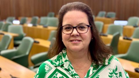 Woman with glasses and shoulder length brunette hair. Wearing a white and blouse with a flower pattern