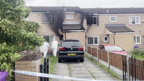 Two terraced houses with visible smoke damage on the brickwork and windows. Two forensic investigators in white overalls approach the front door of the house on the left, which is cordoned off with blue and white police tape. 