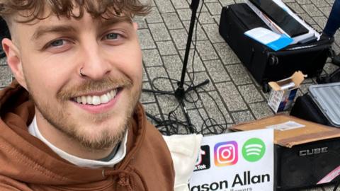A man with brown hair and a small beard, with a black nose ring, wearing a brown hoodie smiles at the camera. He is standing next to music equipment.