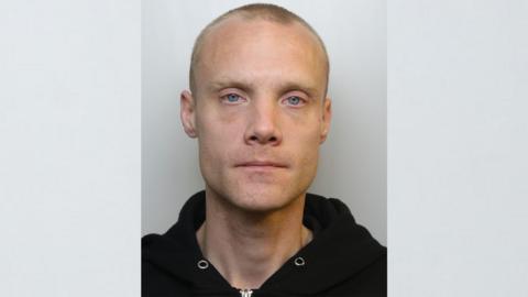 Custody photograph of Mitchell Lee Rogers. He has blonde hair that is shaven, blue eyes and a large visible scar on his left cheek. He is wearing a black hooded jumper with the hood down.