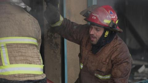 Civil Defence worker fights a fire. He is wearing a red helmet and looks breathless. 