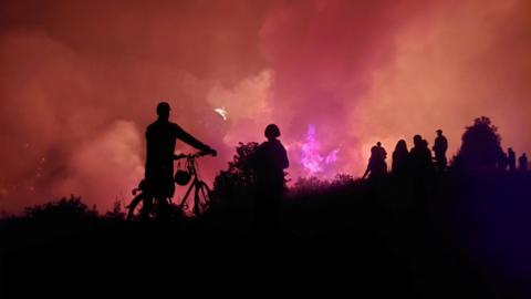 a dark picture of people on a hillside with a fire in the background