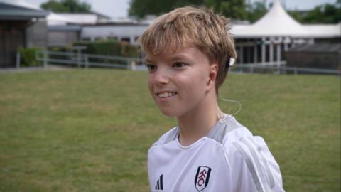 A boy with blond hair, wearing a white football jersey. He is standing on a football pitch at a school. 