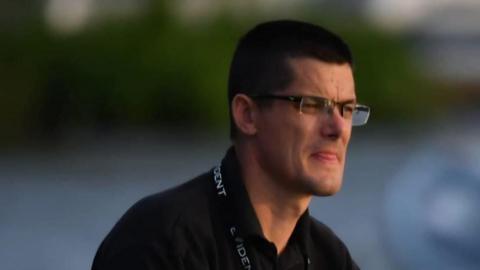 Brett Duncan looks away from the camera. He has short dark hair and wears glasses with a black polo top. 