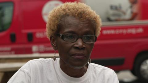 A woman, wearing glasses and earrings, with short blonde hair. She is wearing a white shirt. There is a red van behind her. 