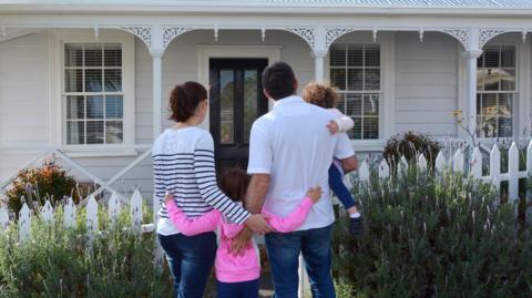 A couple dressed in blue jeans and white shirts. They are standing outside a charming home with intricate white coving, a picket fence and lavender bushes. They are hugging two young girls while looking at the front door of the house.