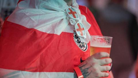 The torso of person wearing an England shirt, flag and St George's Cross glasses with a beer in their hand.