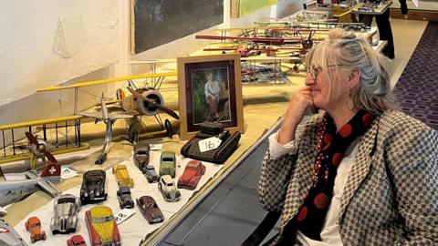 Laurie Stone Wisdom, sat smiling at a photograph of her late husband on a table next to vintage toy cars and model aircraft they collected for more than 30 years