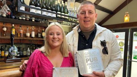 Flor Pearce in a pink top and Joe Buckley in a black shirt and white jacket holding awards