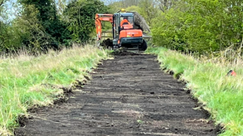 A digger is lifting the top soil and creating a dirt path 