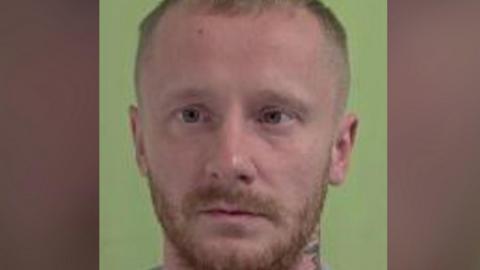 A police mugshot of Fayle, who has short light brown hair and a close-cropped beard and a tattoo on his neck. He is standing against a green background.