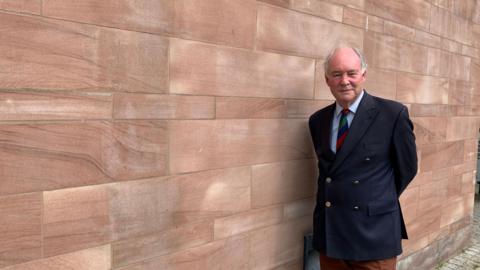 The image shows a late middle-aged man with a balding head and grey hair at the sides, wearing a navy blue blazer over a light blue shirt with a green, blue and red striped tie, standing next to a wall of light red sandstone.