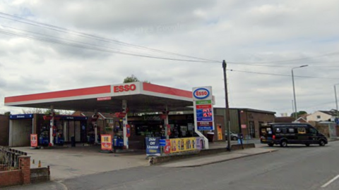 A picture of a petrol station forecourt with the word Esso above the station with a wall to the left of the forecourt and a brick building to the right of the petrol station