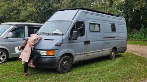 A woman rests her head on the bonnet of a grey van.