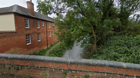 A view of the canal from a bridge with a red-brick hall to the left and trees to the right