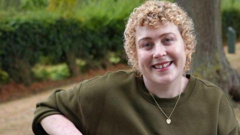 Laura is sitting on a bench in a park. Behind her are a row of trees and some hedges and grass. She is wearing a khaki sweatshirt and has short, curly blonde hair.