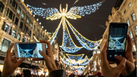 Two sets of hands holding up phones to take photos of the Angel shaped Christmas lights on Regent Street. White curved buildings line each side of the street.