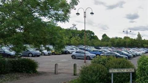 A park and ride car park filled with cars, A street sign in the foreground reads Ten Perch Road.