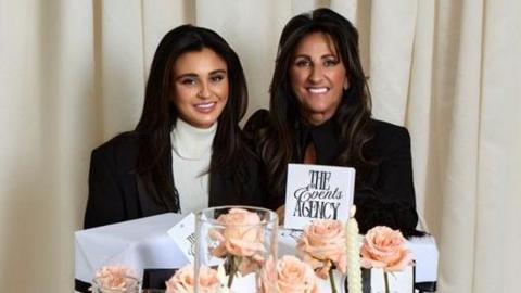 Harriet and Angela Maguire are sitting in front of a table that has been decorated with peach-coloured flowers and candles. There is a white sign which reads: The Events Agency in black lettering.